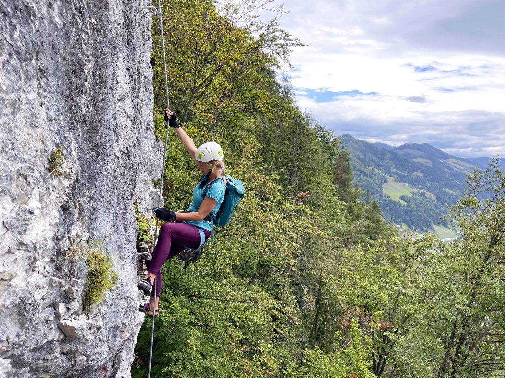 A climber on the via ferrata ascends a steep rock face, secured by a steel cable and steps.