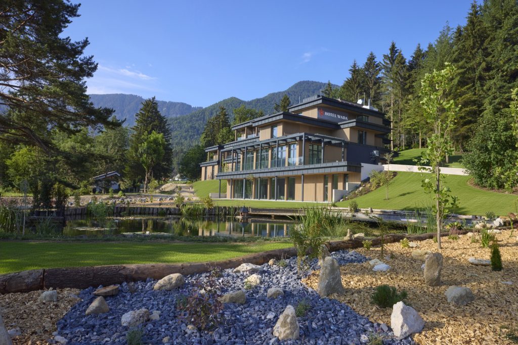 A modern hotel surrounded by nature, with a lake in the foreground, well-maintained greenery, and forested slopes in the background.