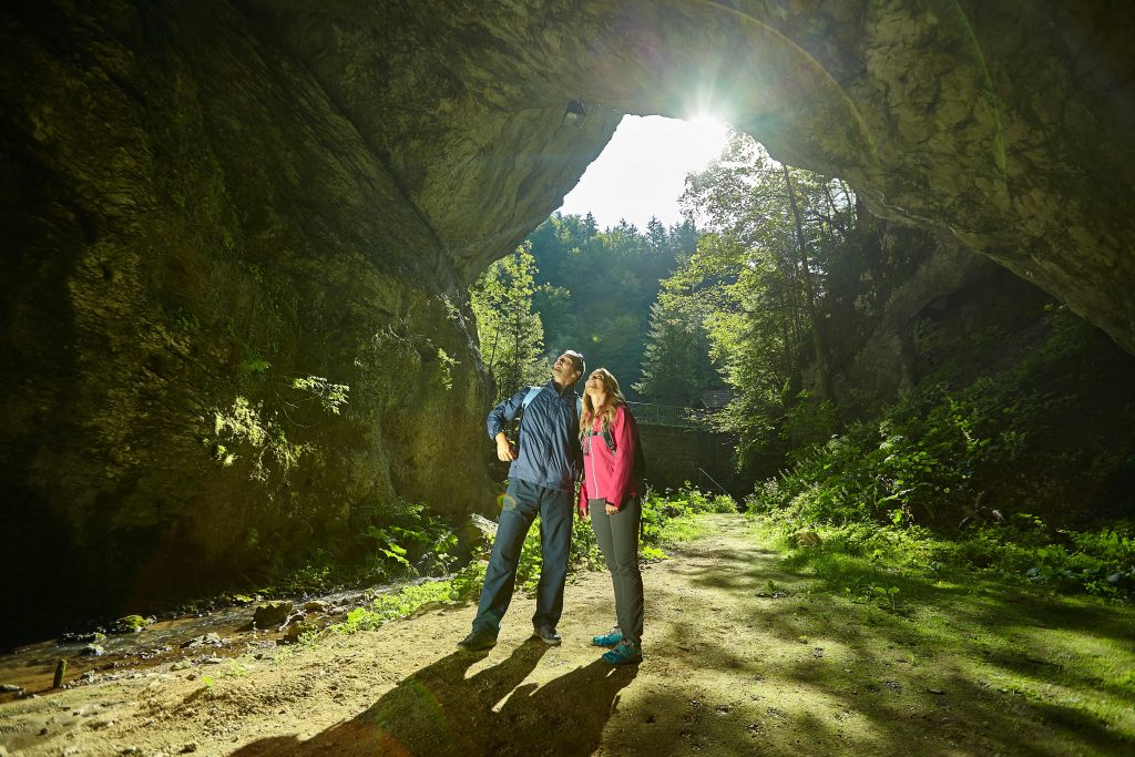 Two hikers explore the scenic entrance to the Huda Luknja cave, where towering rock walls and lush greenery meet.
