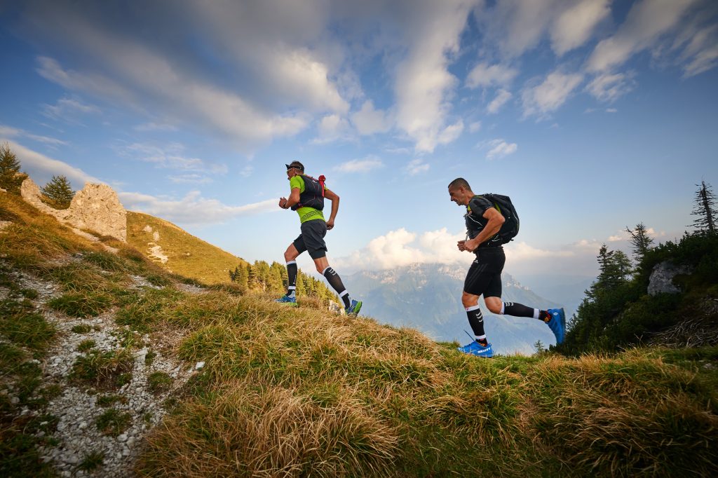 Two runners tackle a varied mountain trail between meadows and forests.