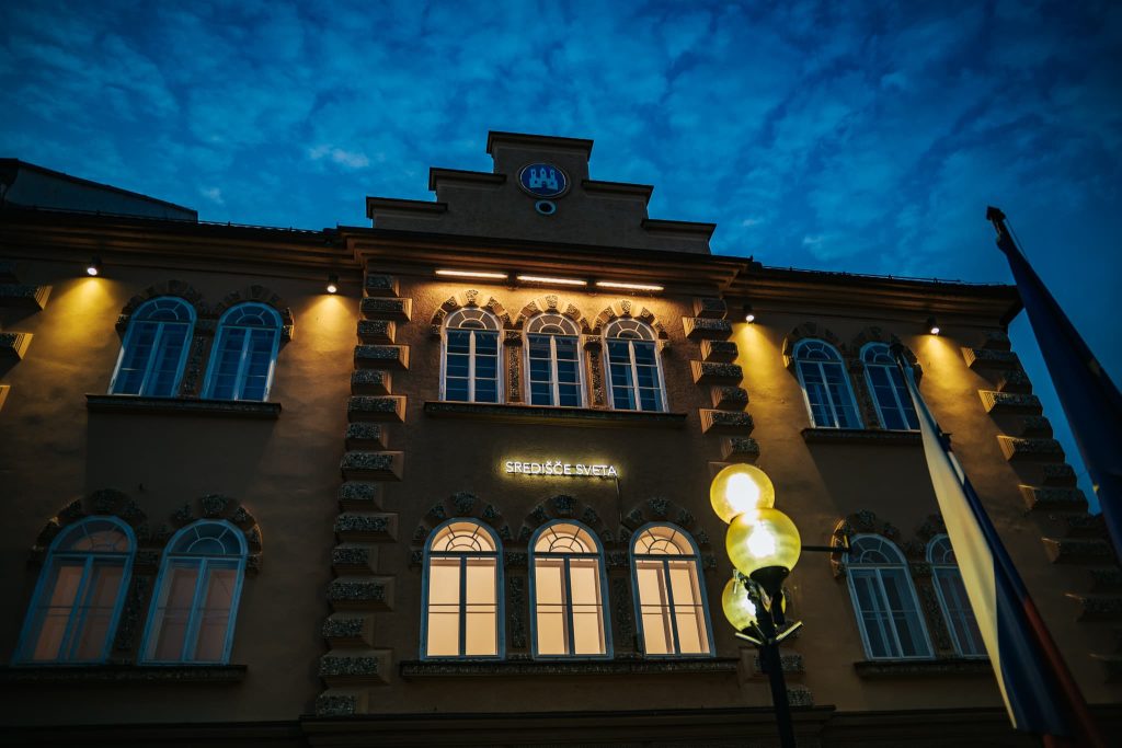 The Koroška Gallery of Fine Arts at night, with its characteristic "Center of the World" inscription, emphasizes its role as a meeting place for art, creativity, and people.