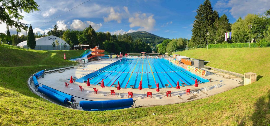 View of the outdoor swimming pool in Ravne na Koroškem with a large swimming pool, water slides and green surroundings.