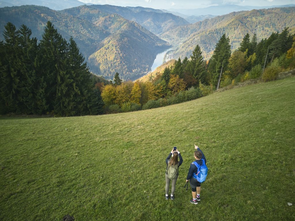 Two hikers admire a wide view of the Drava Valley, forested slopes, and the picturesque Carinthian landscape.