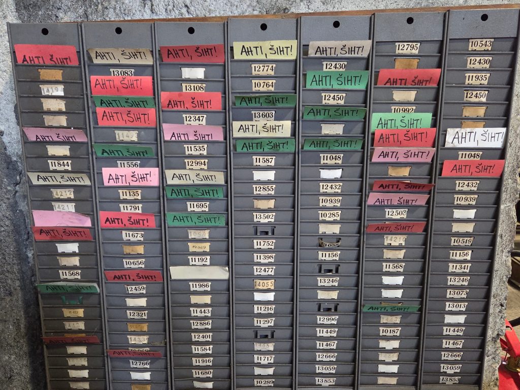 Exhibition display of the former shift time-stamping system with schedules or worker cards and the inscription Ahti, šiht!
