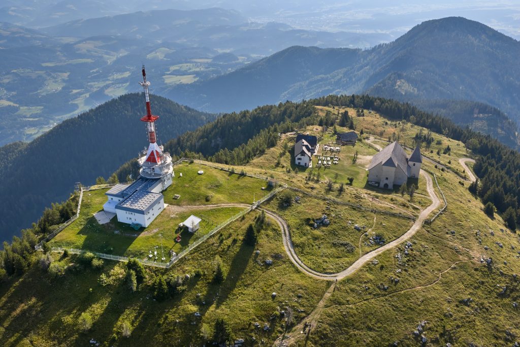 An aerial view of Uršlja Gora with its characteristic transmitter, mountain hut, and the Church of St. Ursula. 