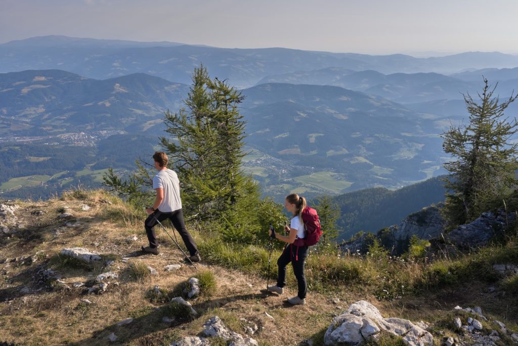 Two hikers on Mount Uršlja Gora with views of Koroška’s varied landscape and valleys deep below them.